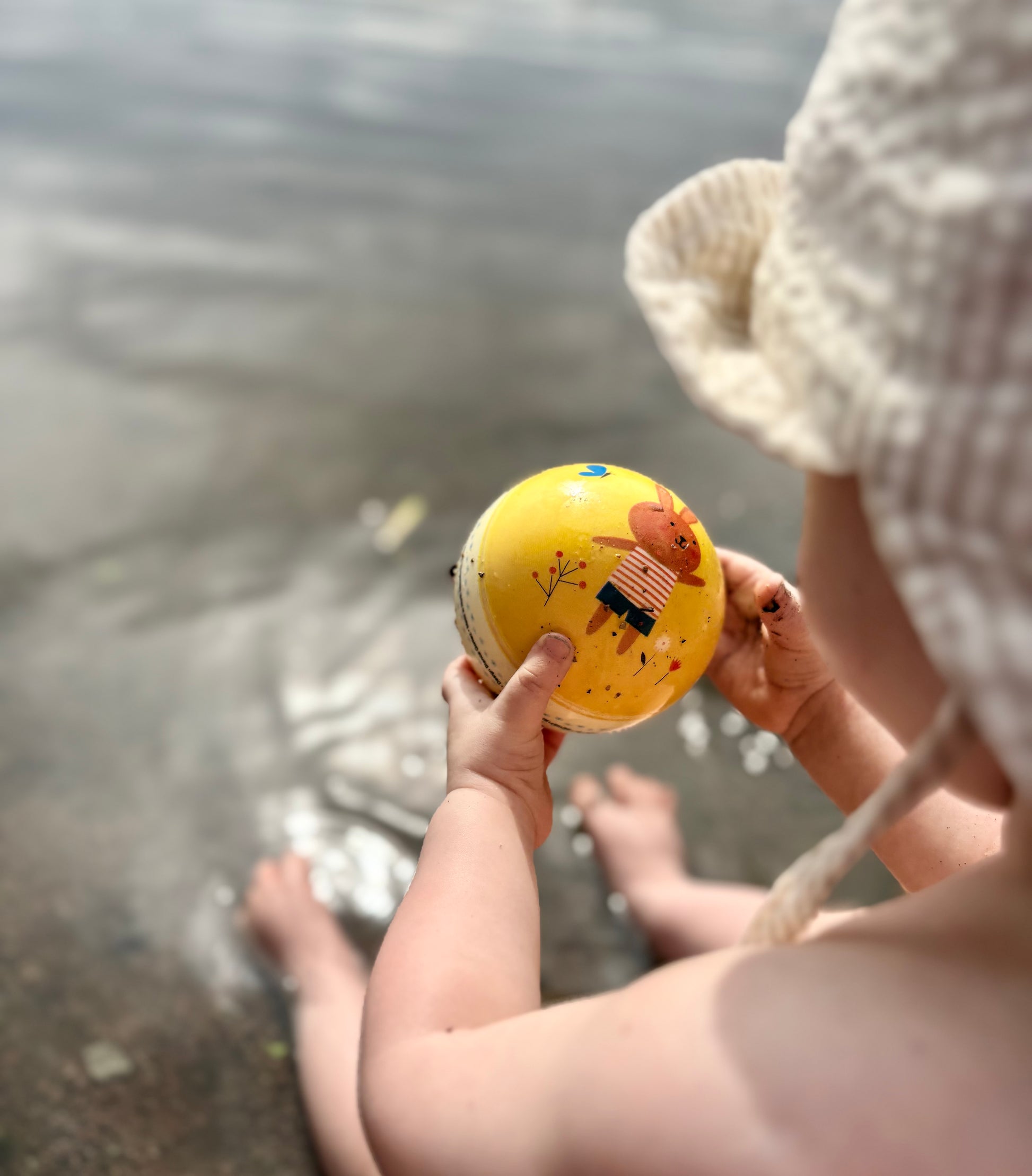 Bebis sitter och tittar på en gul boll sittandes i vattnet på en strand i Sverige