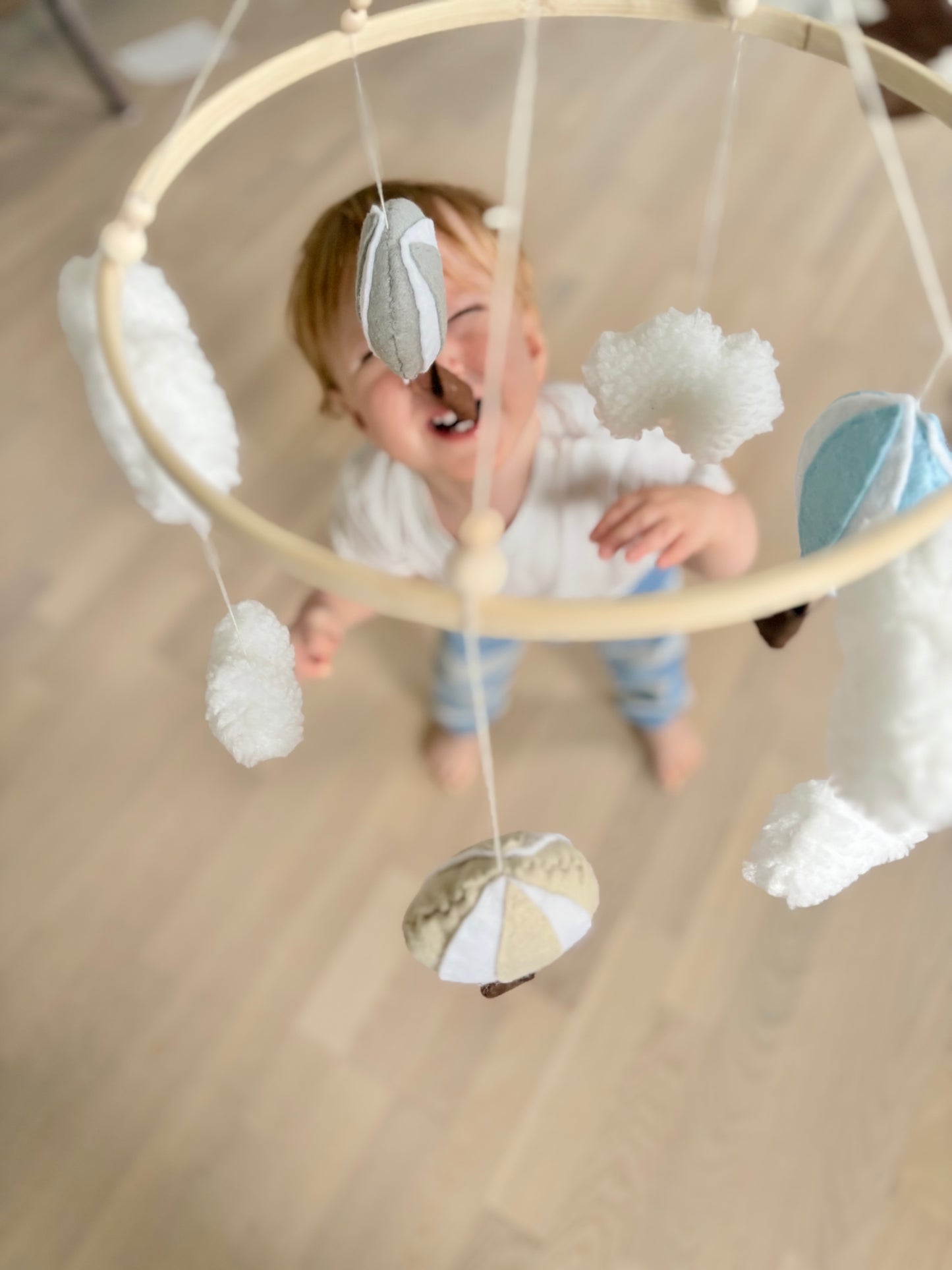 Babymobil i filt – luftballonger bland moln i blå, grå och beige färger svävar över ett litet barn som tittar och skrattar