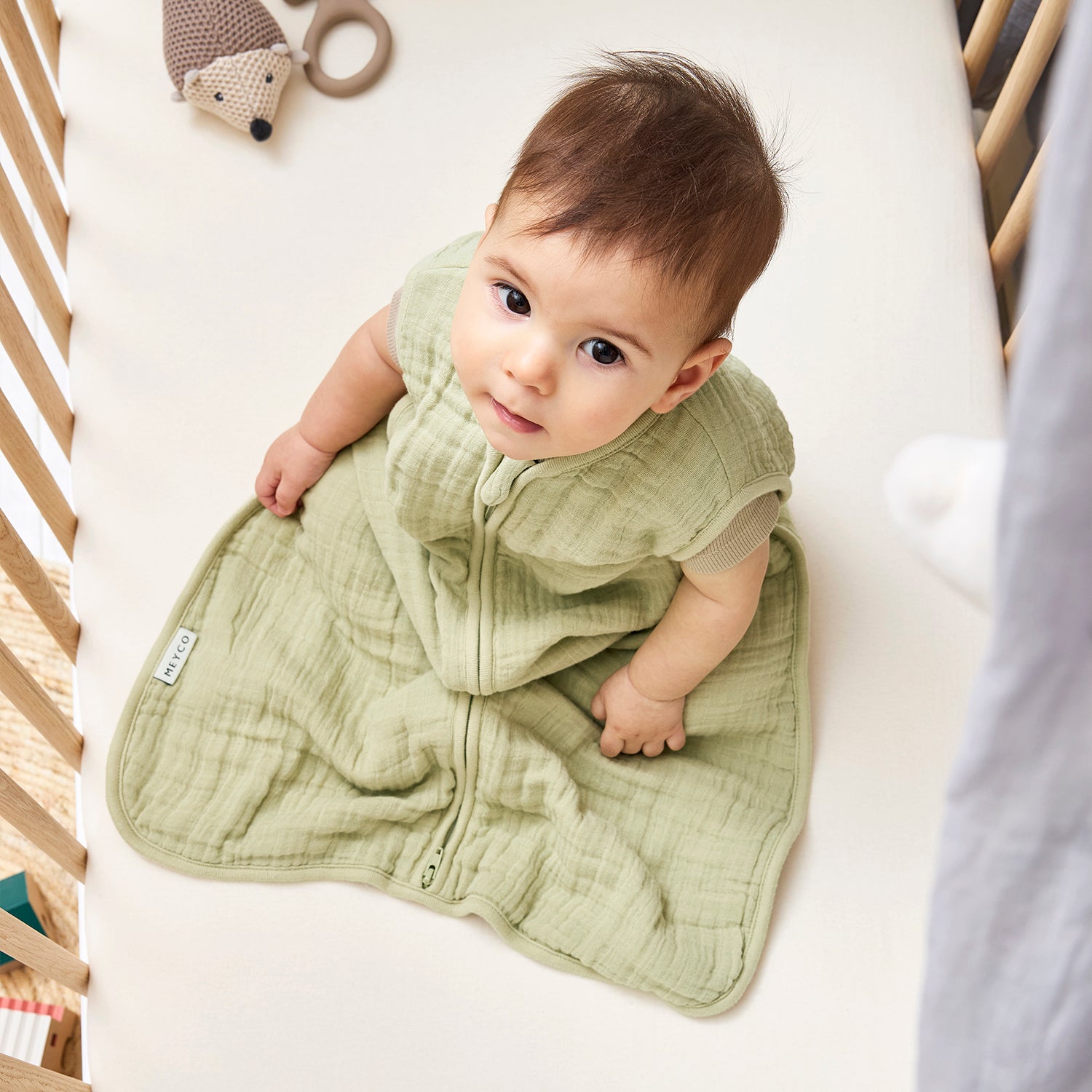 Mjuk sovpåse i muslin för baby – grön färg som ger trygg sömn. Bebis sitter i spjälsäng och tittar upp mot kemeran.